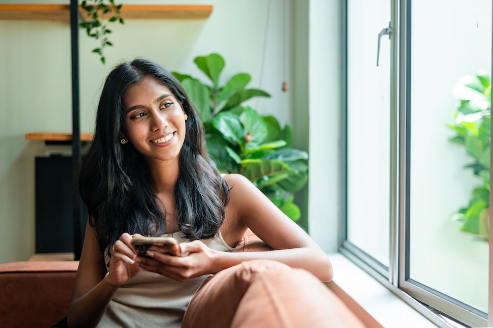 Woman relaxing on sofa, using smartphone and looking away with smile